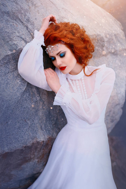 Redhead princess in a white vintage dress lying on the stones. Luxurious outfit with air sleeves. Sleeping Beauty. Creative blue makeup, silver tiara. Art processing portrait photography - Fotó, kép