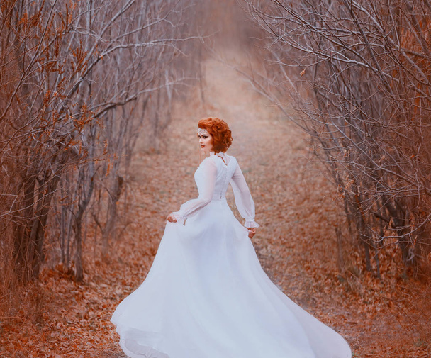 The redhead princess in a white vintage dress runs in a tunnel from the trees and looks back. Luxurious outfit with a long train flutters in the wind. Sleeping nature, golden autumn, warm colors - Photo, Image