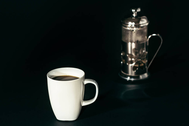 eine Tasse Kaffee mit einem verschwommenen Kolben - französische Kaffeemaschine vor schwarzem Hintergrund. - Foto, Bild