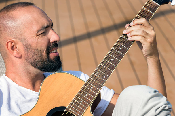 あごひげと短いバズヘアを持つスペイン人男性の肖像画、 屋外でアコースティックギターを演奏. - 写真・画像