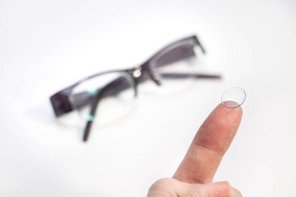 ein männlicher Finger, der eine Kontaktlinse über einer verschwommenen Brille als Hintergrund hält. - Foto, Bild