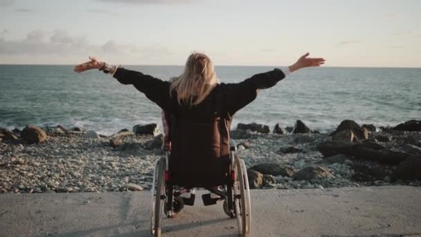 Alone disabled woman is sitting in wheelchair and holding hands up on sea coast - Footage, Video