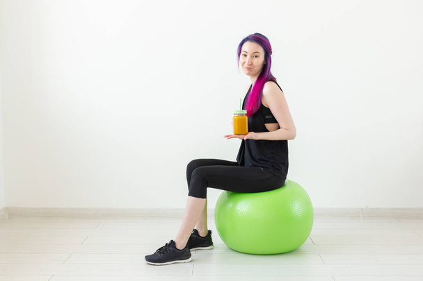 Χαριτωμένο νέος μικτός αγώνας χίππη κορίτσι με χρωματιστά μαλλιά κάθονται σε ένα πράσινο γυμναστήριο και κρατώντας ένα χυμό πρωτεΐνης μπανάνα στα χέρια της σε λευκό φόντο. Υγιεινή διατροφή και αίσθηση άσκησης. Χώρος αντιγραφής - Φωτογραφία, εικόνα