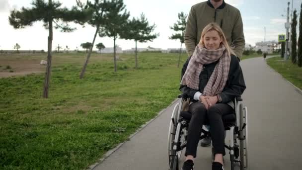 Mujer enferma joven está sentado en silla de ruedas, el hombre la está ayudando y rodando
 - Metraje, vídeo