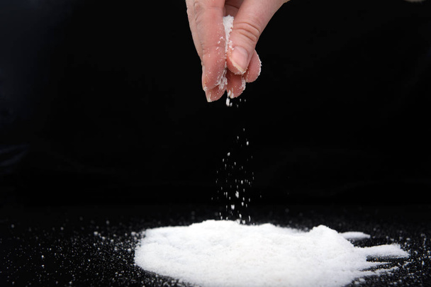 in de vingers van een jonge vrouw een snufje zout, dat ze op een heuvel zout giet. Afbeelding op zwarte achtergrond. Concept-zoutloos dieet. - Foto, afbeelding