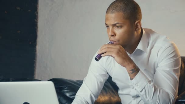 selbstbewusst gut gekleideter afrikanisch-amerikanischer Geschäftsmann benutzt Laptop und dampft in seinem Büro eine elektronische Zigarette, während er auf dem Sofa sitzt - Filmmaterial, Video