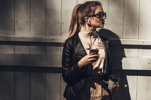 Portrait of Stylish Girl Wears Women Overalls and Black Leather Jacket Drink Coffee, Using Smartphone - Photo, Image