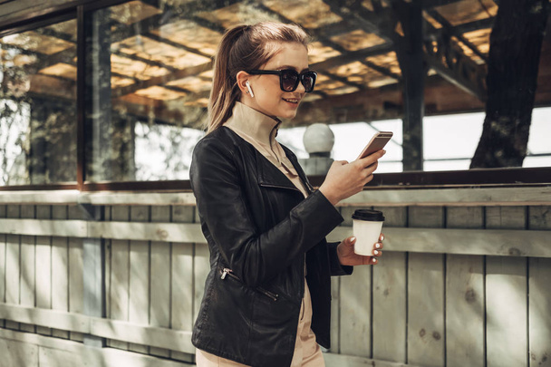 Retrato de chica con estilo lleva monos de mujer y chaqueta de cuero negro Beber café, Usando Smartphone
 - Foto, Imagen