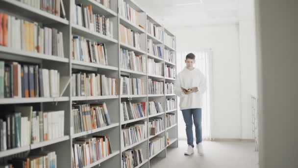 Przystojny młody student spacerujący między półką książki z książką w rękach - Materiał filmowy, wideo