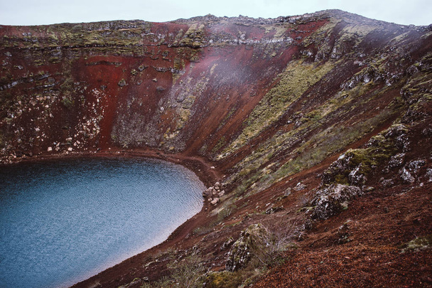 Colinas vermelhas carmesim de uma cratera vulcânica que rodeia as águas azuis do lago Kerith (Kerid) na Islândia
 - Foto, Imagem