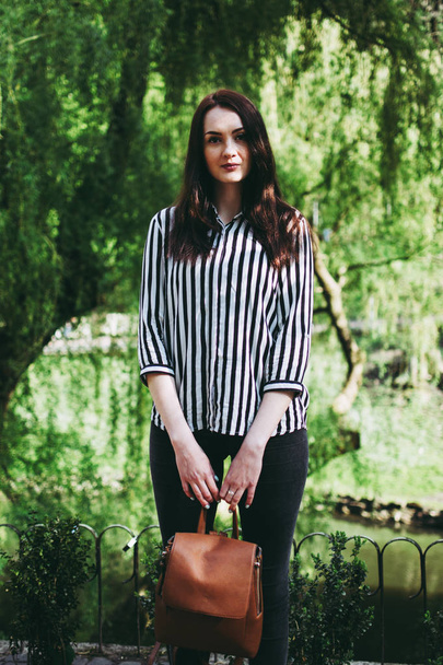 Portrait de jolie jeune brune dans un parc. Chemise élégante rayée noire et blanche. Modèle étudiant tenant un sac marron
 - Photo, image