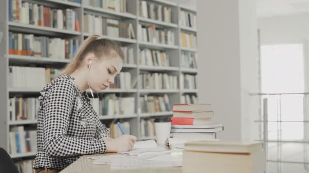 Koncentrált lány hallgató fülhallgatóval készül vizsga és írás jegyzetek, miközben ül asztalnál az egyetemi könyvtárban - Felvétel, videó