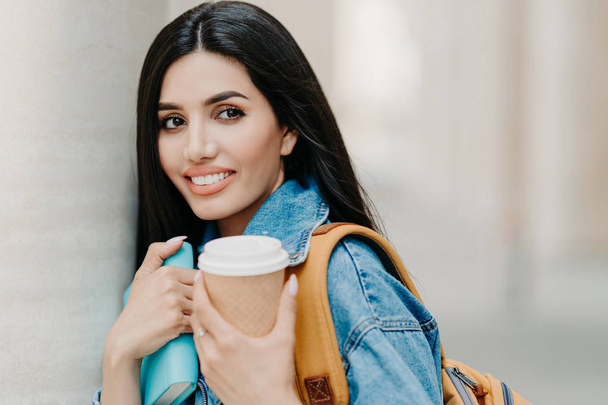 Outdoor shot of brunette female student with makeup, dark long hair, dressed in denim jacket, holds takeaway coffee, textbook, carries rucksack, looks somewhere into distance, strolls in city - Photo, image