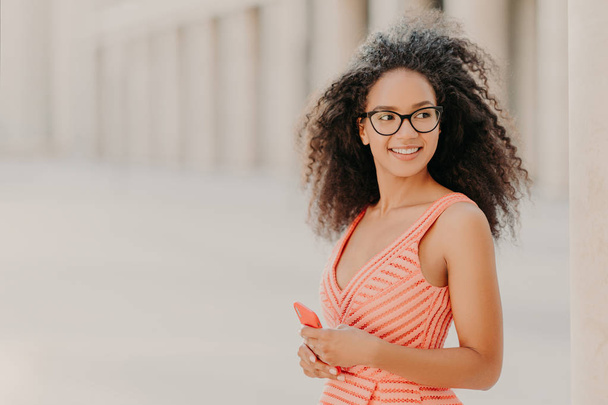 Pleased millennial girl with curly hair, wears pink dress, optical glasses, focused aside with smile, stands outdoor, waits for call, enjoys high speed internet connection, updates new application - Photo, Image