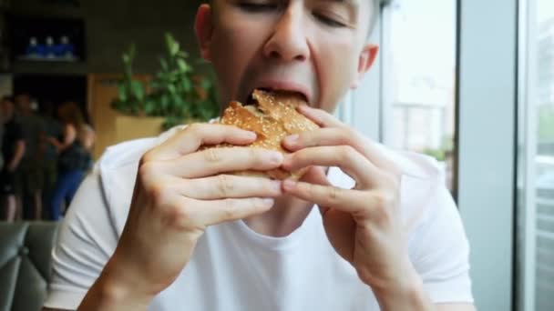 Zbliżenie młodego głodnego mężczyzny zjada Hamburger w kawiarni, restauracji szybkiej żywności - Materiał filmowy, wideo