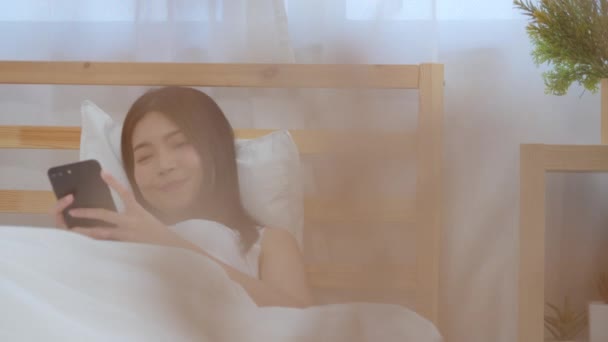 Pomalý pohyb-mladá asijská žena, která používá smartphone a kontroluje společenské sdělovací prostředky, se šťastně usmívá, když leží na posteli, když se ráno probudila, atraktivní Japonka se usmívá v ložnici doma. - Záběry, video