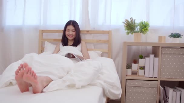 Beautiful attractive young Asian woman reading a book while lying on the bed when relax in her bedroom at home in the morning. Lifestyle women using relax time at home concept. - Imágenes, Vídeo