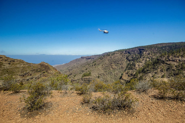 Πέτρινο μονοπάτι που περιβάλλεται από πεύκα την ηλιόλουστη μέρα. Καθαρός μπλε ουρανός και σύννεφα κατά μήκος της γραμμής του ορίζοντα. Δρόμος σε ξηρά ορεινή περιοχή με ξύλα φύλλων βελόνας. Ελικόπτερο που πετάει πάνω από το φαράγγι. Τενερίφη - Φωτογραφία, εικόνα