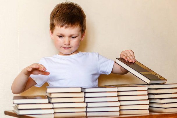 een schattige zes-jarige jongen zit aan een bureau en zet boeken op stapels. - Foto, afbeelding