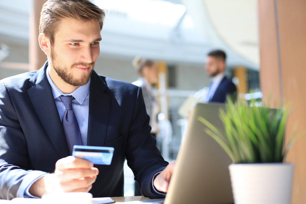 Hombre sonriente sentado en la oficina y paga con tarjeta de crédito con su computadora portátil. - Foto, Imagen