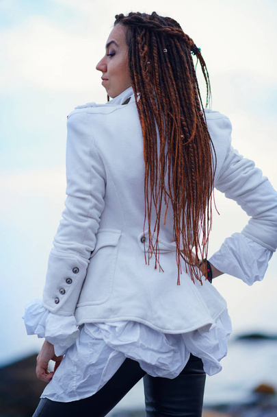 дредлоки модна дівчина, одягнена в білий піджак і чорні шкіряні штани позують біля моря ввечері
 - Фото, зображення