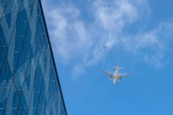 Авиаплан оставляет след в голубом небе. Авиалайнер взлетает над зданием. Белое облако в голубом небе, в котором летит самолет
 - Фото, изображение