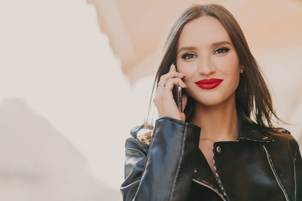 Attractive lovely female enjoys tariffs in roaming, talks on mobile phone with best friend, dressed in fashionable clothes, wears red lipstick, makeup, stands on blurred background of some building - Foto, Imagem