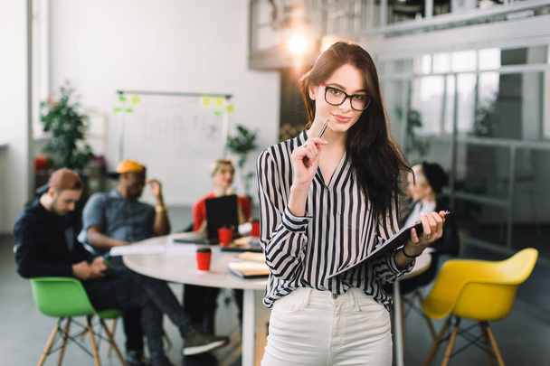 Portrait of a happy, smiling dark haired businesswoman in white pants and shirt, wearing glasses, standing in loft office. Young business people at the table on the background - Фото, зображення