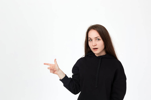 Portrait of a pretty young brunette in a black sweatshirt on a white background pointing with fingers to the left. - Foto, immagini