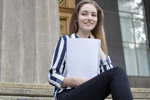 schöne Studentin sitzt auf den Stufen in der Nähe des College mit einem Rucksack, hält Papiere in den Händen und schaut auf, sie lernt Lektionen in der Pause - Foto, Bild