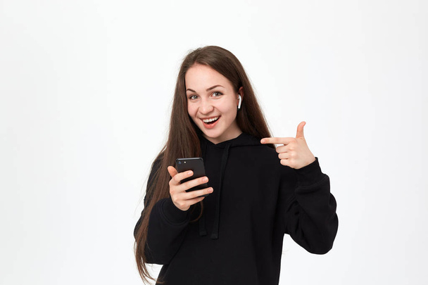 Studio shot of a beautiful young brunette woman with wireless headset and phone looking happy at the camera and pointing at the phone screen while standing over white background. - Valokuva, kuva