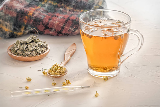 Chá de ervas com camomila farmacêutica, cachecol quente e termômetro médico em uma mesa branca
 - Foto, Imagem