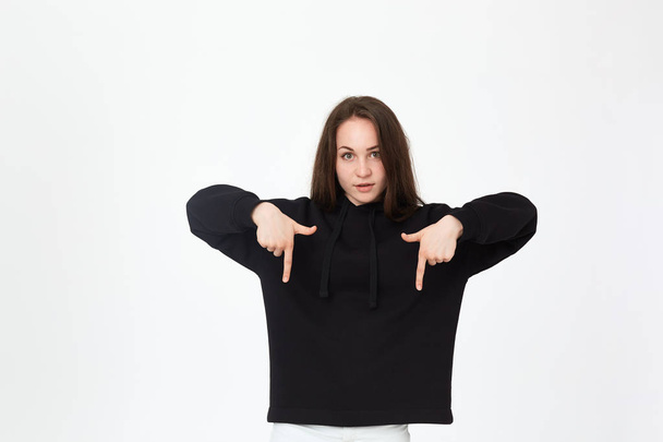 Portrait of a pretty young brunette in a black sweatshirt on a white background pointing down with fingers. - Fotografie, Obrázek