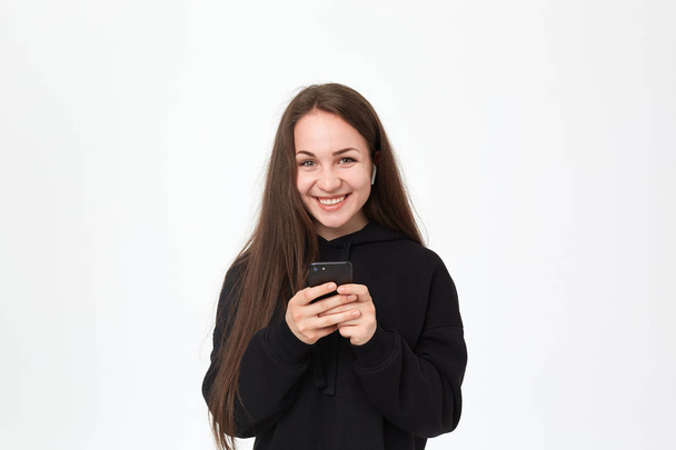Beyaz arka plan üzerinde dururken kablosuz kulaklık ve telefon ile kamera mutlu görünümlü güzel bir genç esmer kadın Stüdyo çekim. - Fotoğraf, Görsel