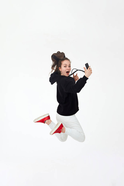 Młoda ładna brunetka dziewczyna w czarnym kapturze, białe spodnie i czerwone trampki trzymać telefon komórkowy i skacze izolowane na białym tle. - Zdjęcie, obraz