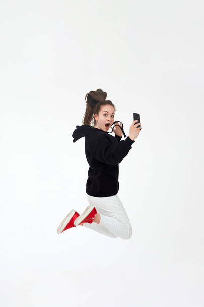 Joven chica morena bonita en sudadera con capucha negra, pantalones blancos y zapatillas rojas sostienen el teléfono móvil y salta aislado sobre fondo blanco
. - Foto, imagen