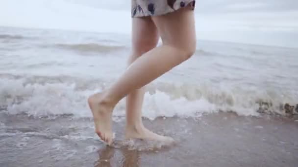 Menina em um vestido curto com flores andando descalço na praia e mar lava os pés. Close-up - Filmagem, Vídeo