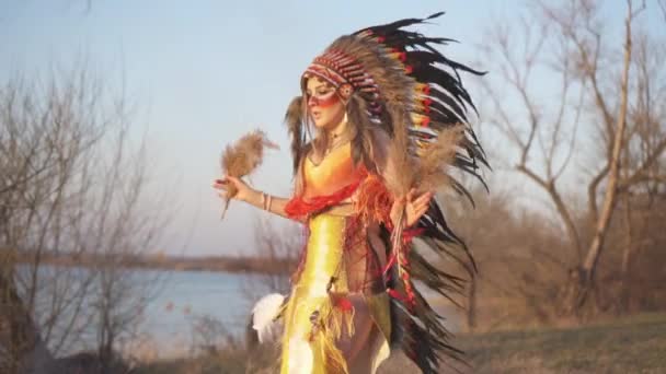 Gyönyörű lány indián fejdíszben és jelmezben, színes sminkkel, ahogy ugrál a rituáléban a naplementében, indiai fűvel a kezében.. - Felvétel, videó