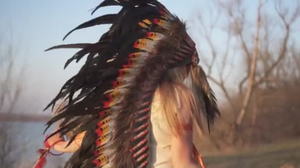 Gyönyörű lány indián fejdíszben és jelmezben, színes sminkkel a naplemente rituáléján, indián fűvel a kezében.. - Felvétel, videó