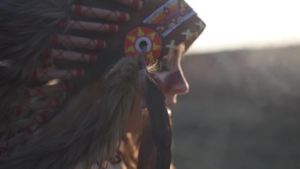 Vista laterale della bella ragazza in indiano nativo americano copricapo e costume con trucco colorato alla luce del tramonto in fumo. Lei sussurra. Primo piano - Filmati, video