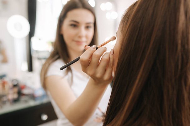 Beautiful femalemakeup artist doing makeup for a young redhead girl in a beauty salon sitting in front of a large mirror. Concept of preparation for the holiday and meeting - Φωτογραφία, εικόνα
