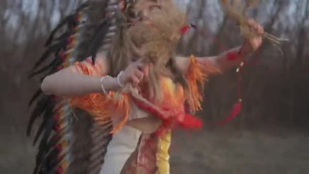 Gyönyörű lány indián fejdísz és jelmez színes smink teszi mozgások indiai fű kezében az arca közelében naplemente fény. A modell lenéz. Sorghastrum dilinyósok. Közelkép - Felvétel, videó