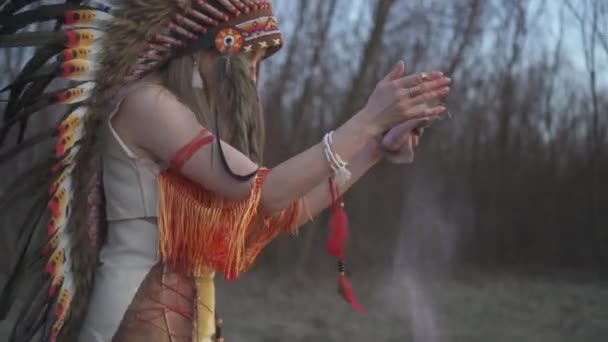 Beautiful girl in native american indian headdress and costume with colorful make-up in the rituale dance on the sunset. She hiting the pouch and pink powder flows from it - Footage, Video