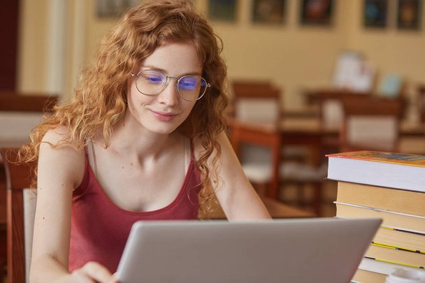 Innenaufnahme einer konzentrierten schönen Studentin, die mit Laptop in der Bibliothek arbeitet, Notizen auf ihrem tragbaren Computer macht und im Lesezimmer fleißig lernt. Hochschule bereitet sich auf Prüfungskonzept vor. - Foto, Bild