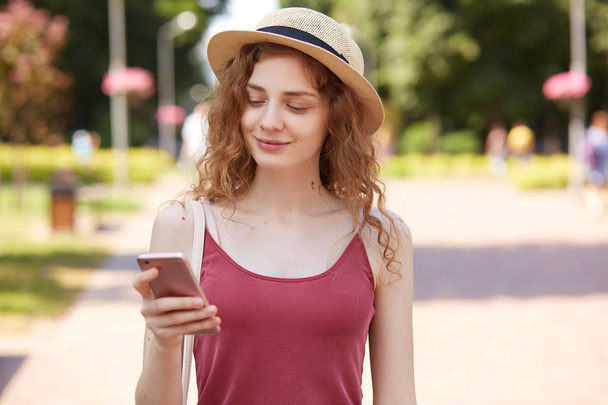 Delighted happy young lady having pleasant facial expression, smiling sincerely, reading news, holding smartphone, using it actively, wearing red shirt and fashionable summer hat. Social medias. - Foto, imagen