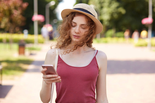 Tender sweet curly haired girl walking along green summer street, having smartphone in her hand, being focused on her mobile phone, making new post attentively, wearing casual clothes. Summer concept. - Zdjęcie, obraz