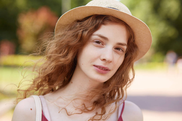 Close up portrait of attractive young lady in straw hat. Foxy haired charming woman with brown eyes looking at camera with gentle, has calm facial expression, posing isolated over park background. - Foto, Bild