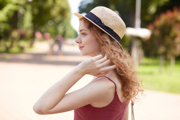 Profile of European romantic attractive young female looking aside, touching her hair with fingers, wearing straw hat and red shirt, being fond of accessories, having sincere smile on her face. - Fotografie, Obrázek