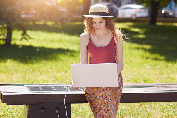 Concentrated pensive young lady sitting in straw hat, holding her white laptop, looking at its screen attentively, charging her device with USB, working during lunchtime outdoors. Youth concept. - 写真・画像
