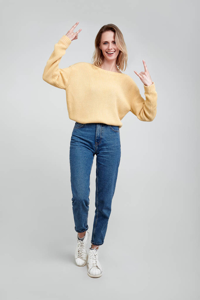 Full length portrait of a young blond woman showing rock gesture with hands isolated over gray background. dressed in casual clothes - Fotó, kép